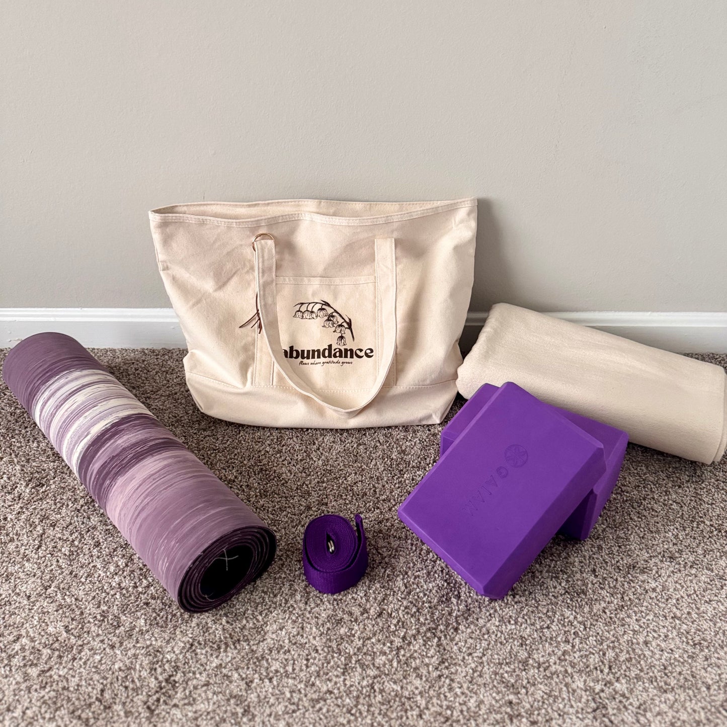 Yoga mat, strap, block, and tote bag on a carpeted floor with a gray wall background.