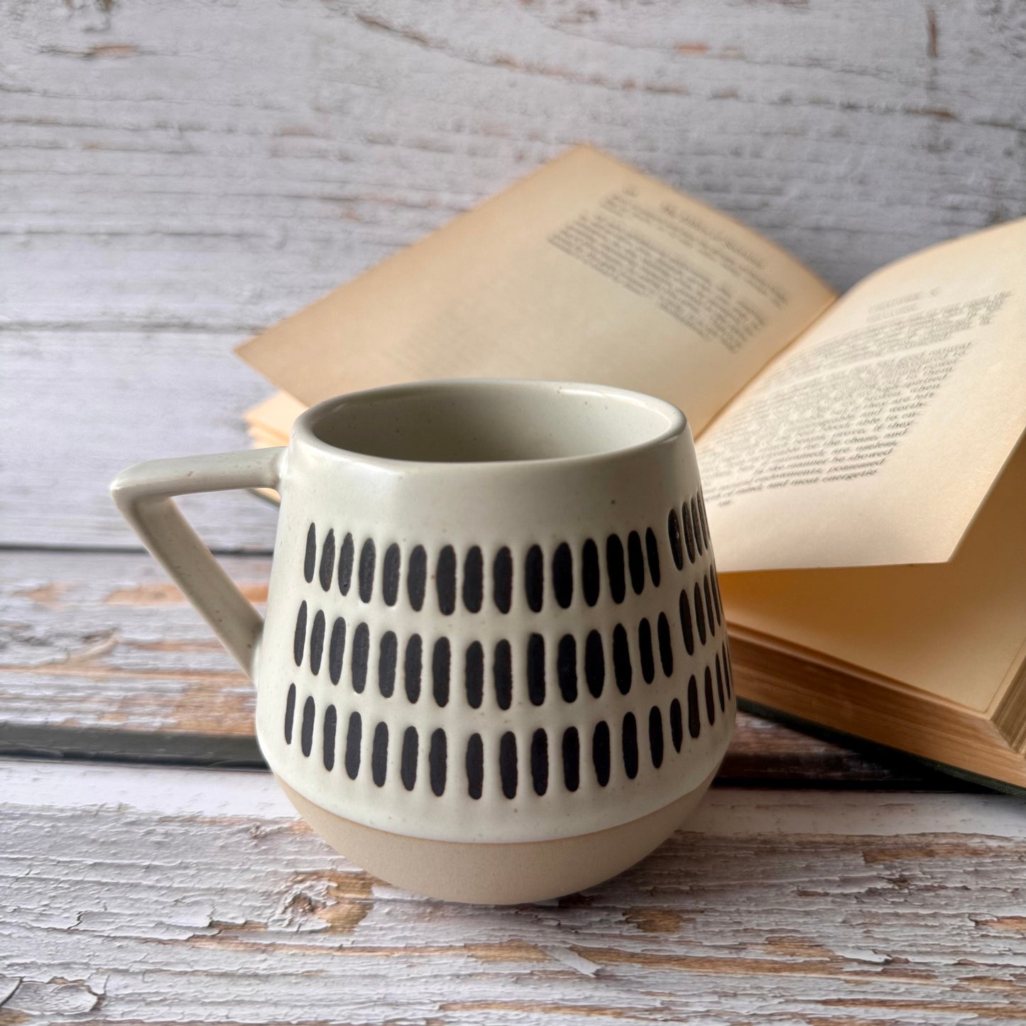 Warm beige and soft white with natural brown undertones ceramic mug with textured design on a wooden surface with an open book in the background