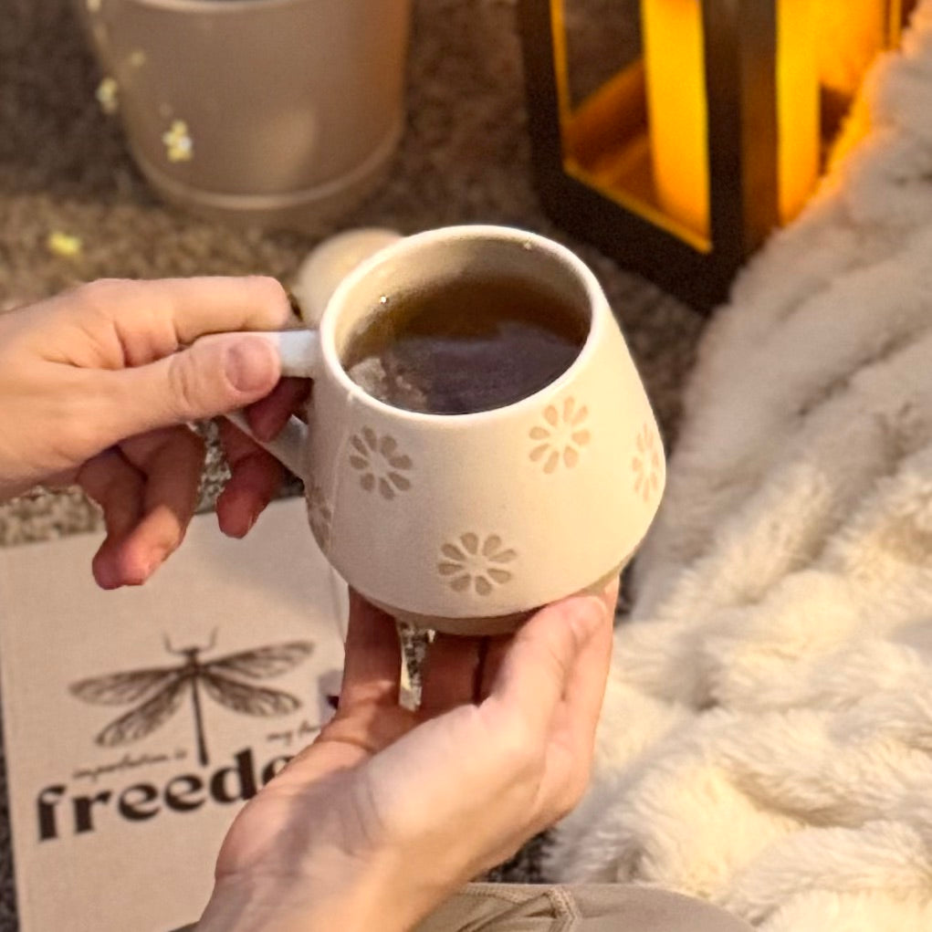 Person holding a decorative warm beige and soft white with natural brown undertones mug with a floral pattern, sitting on a textured surface.