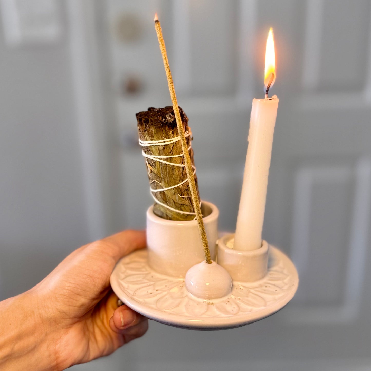 Hand holding a white ceramic holder with incense sticks and a lit candle against a neutral background
