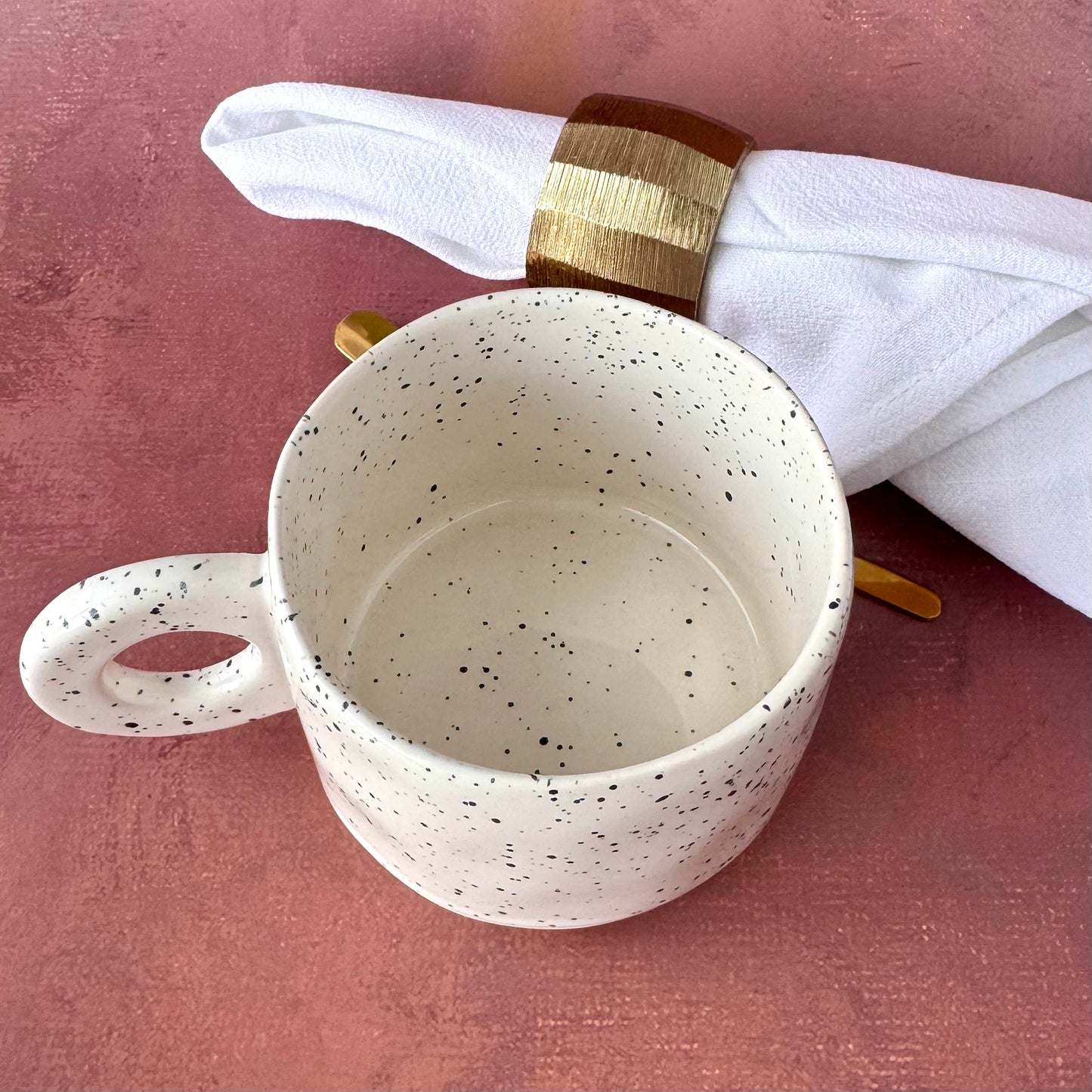 Speckled white ceramic mug with a gold napkin ring on a pink surface