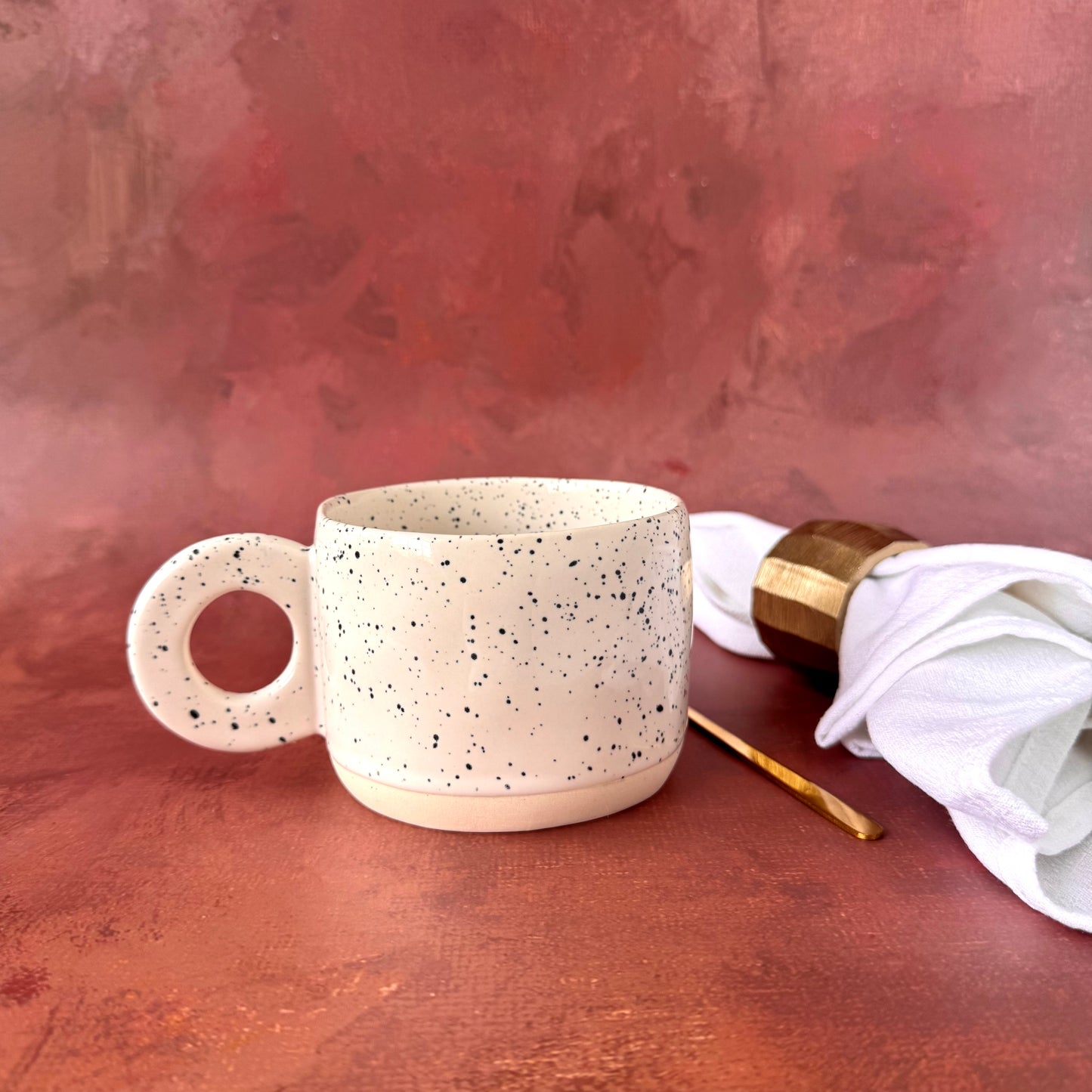 Speckled white ceramic mug on a textured surface with a napkin and gold ring.