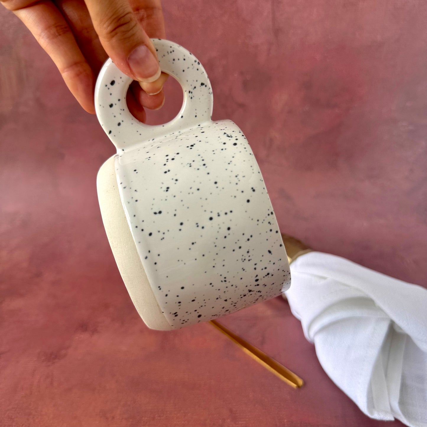 Speckled white stylish ceramic cup held by a hand on a pink background