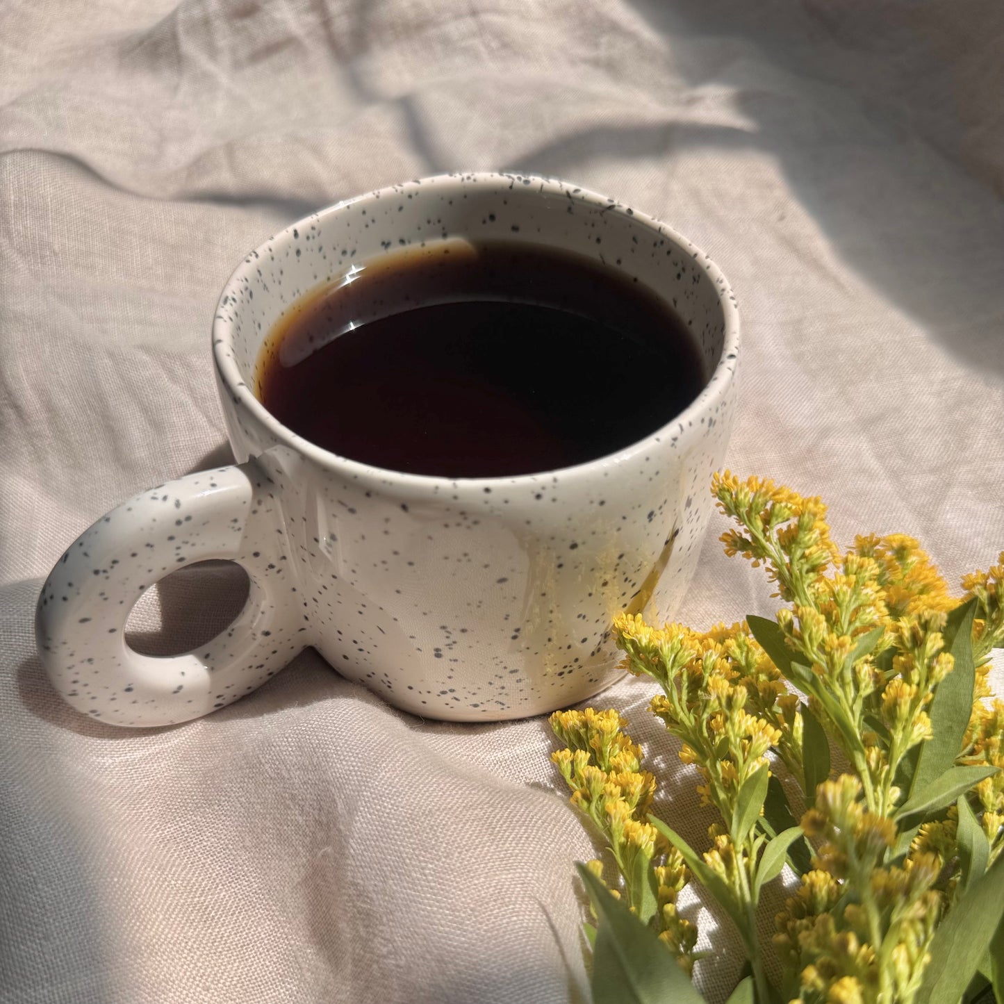 Speckled white stylish ceramic mug with coffee on a light fabric surface with yellow flowers.