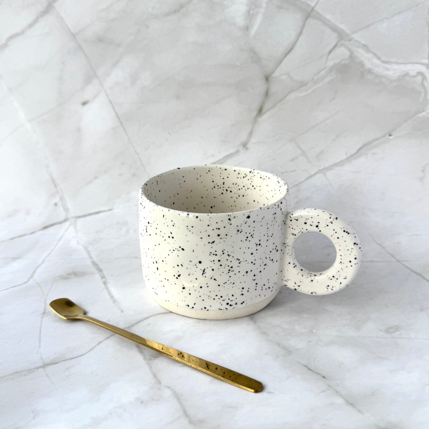 Speckled white stylish ceramic mug with a gold spoon on a marble surface