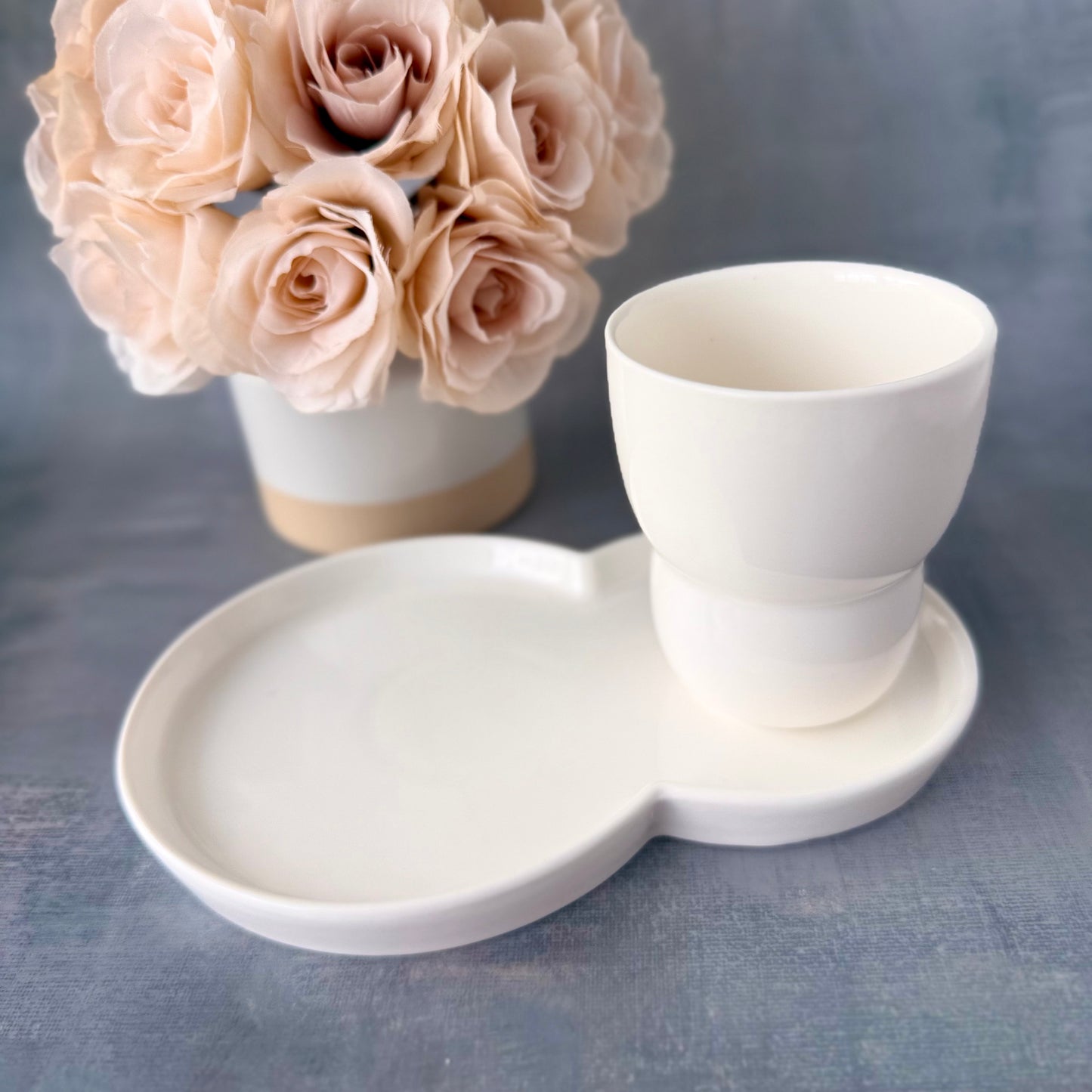 White ceramic cup and tray with a vase of beige roses on a gray background