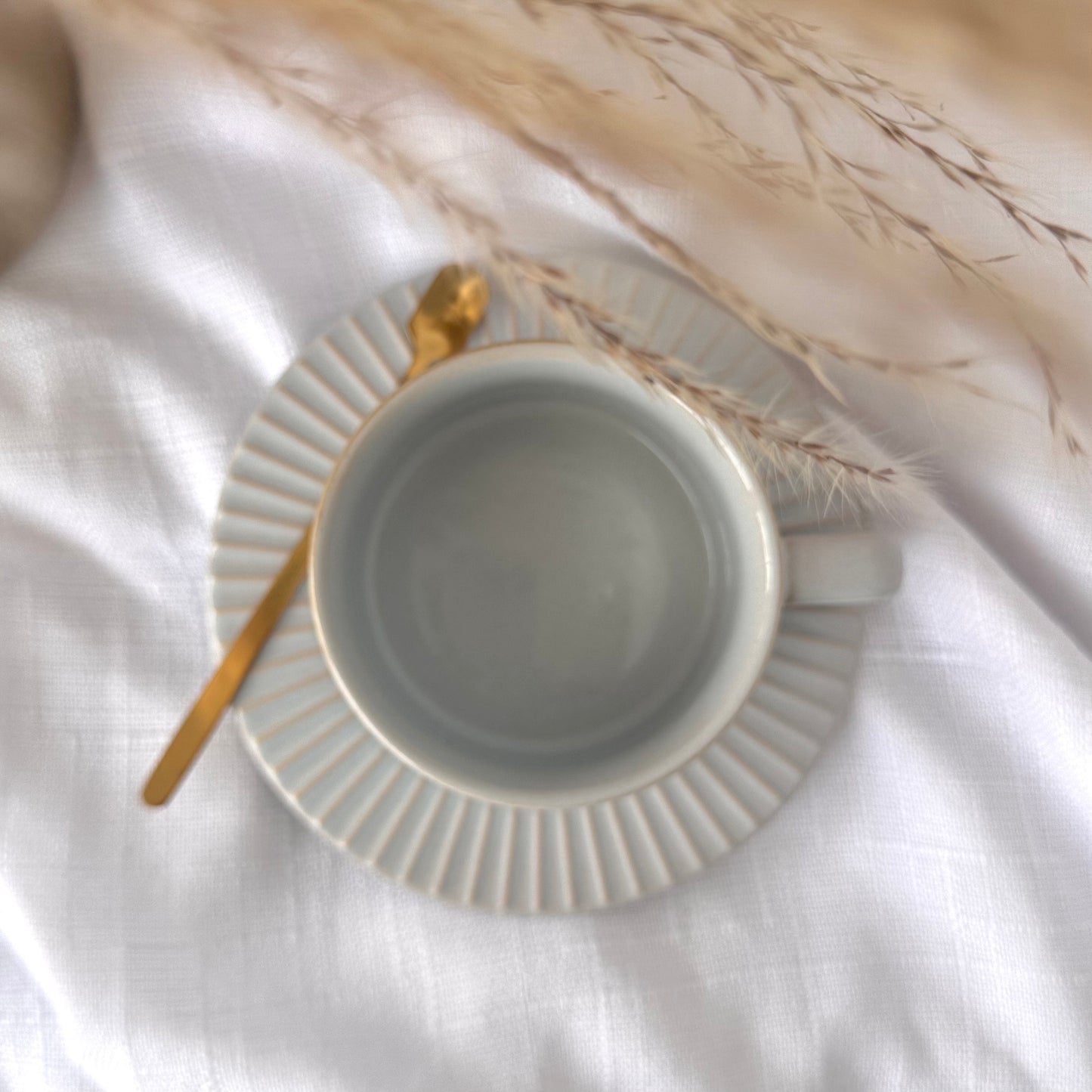 Calming light gray glaze with soft blue undertones ceramic cup with gold spoon on a white surface with dried plants