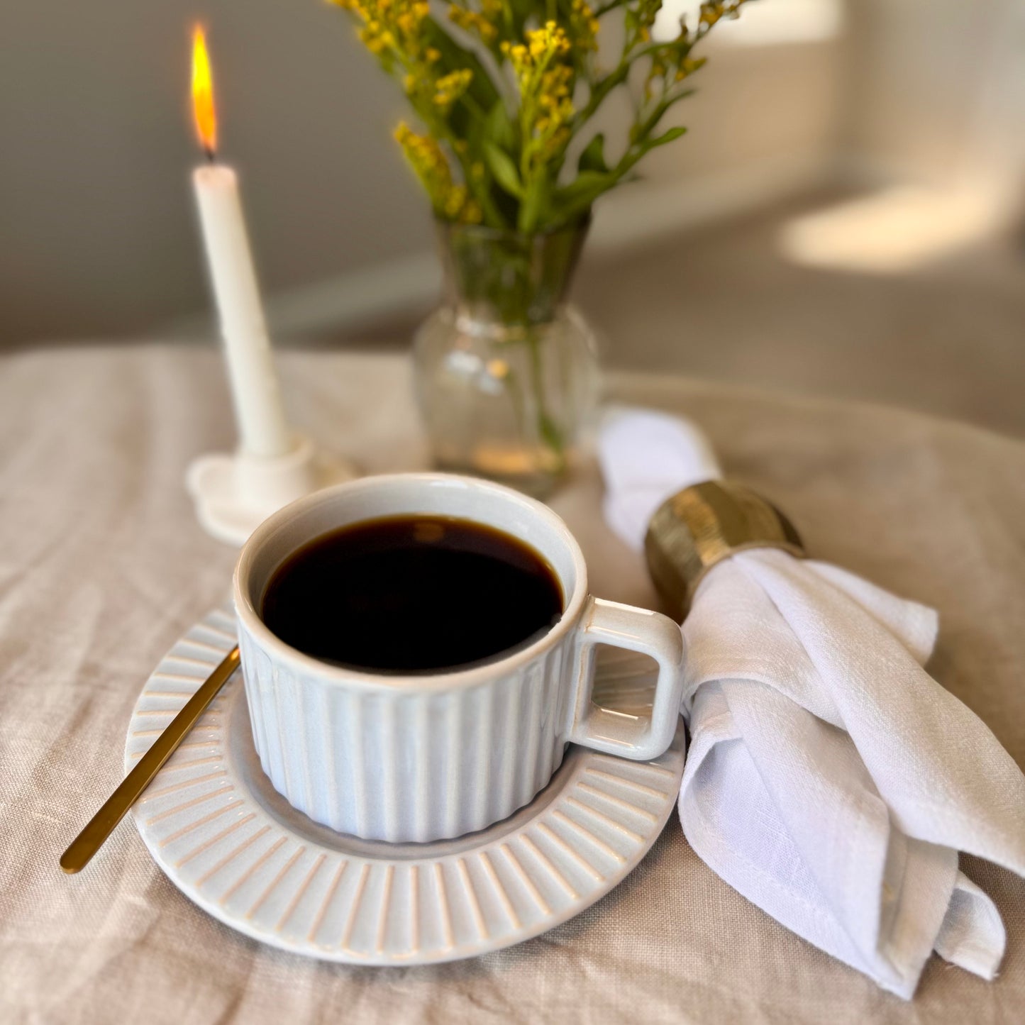 Calming light gray glaze with soft blue undertones mug with black coffee on a beige tablecloth, next to a lit candle and a vase with flowers.