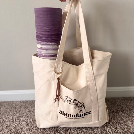 Beige tote bag with 'abundance' text and a purple cup inside, on a neutral background