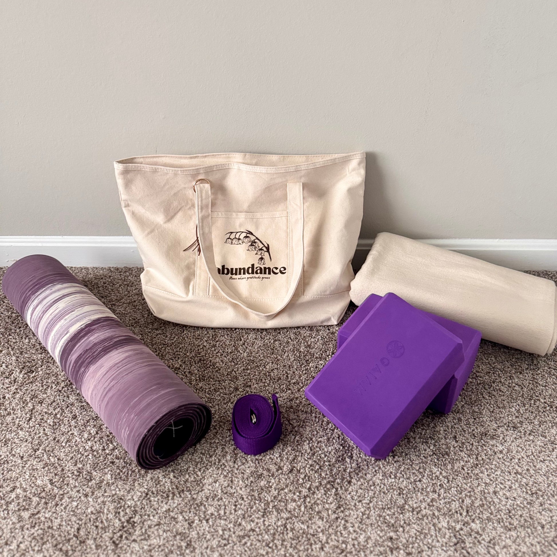 Yoga mat, strap, block, and tote bag on a carpeted floor with a gray wall background.