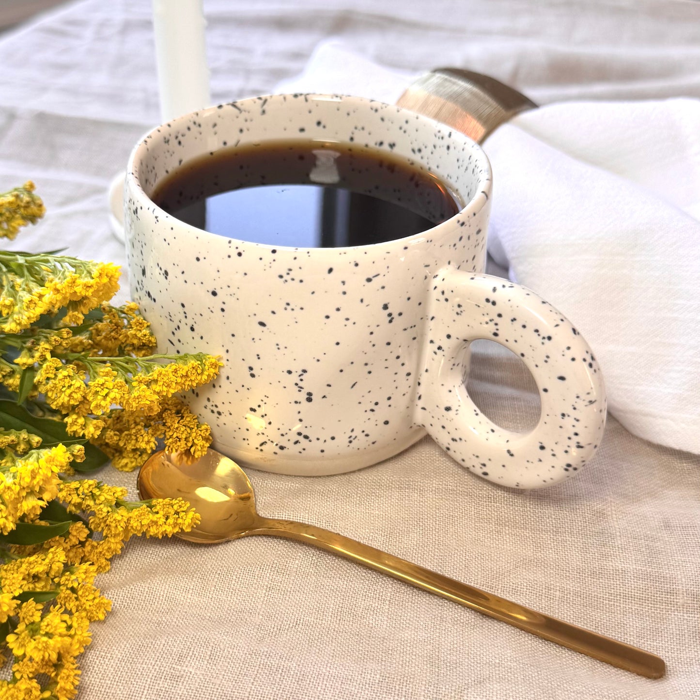 Speckled white ceramic mug with hot coffee, gold spoon, and yellow flowers on a beige surface.