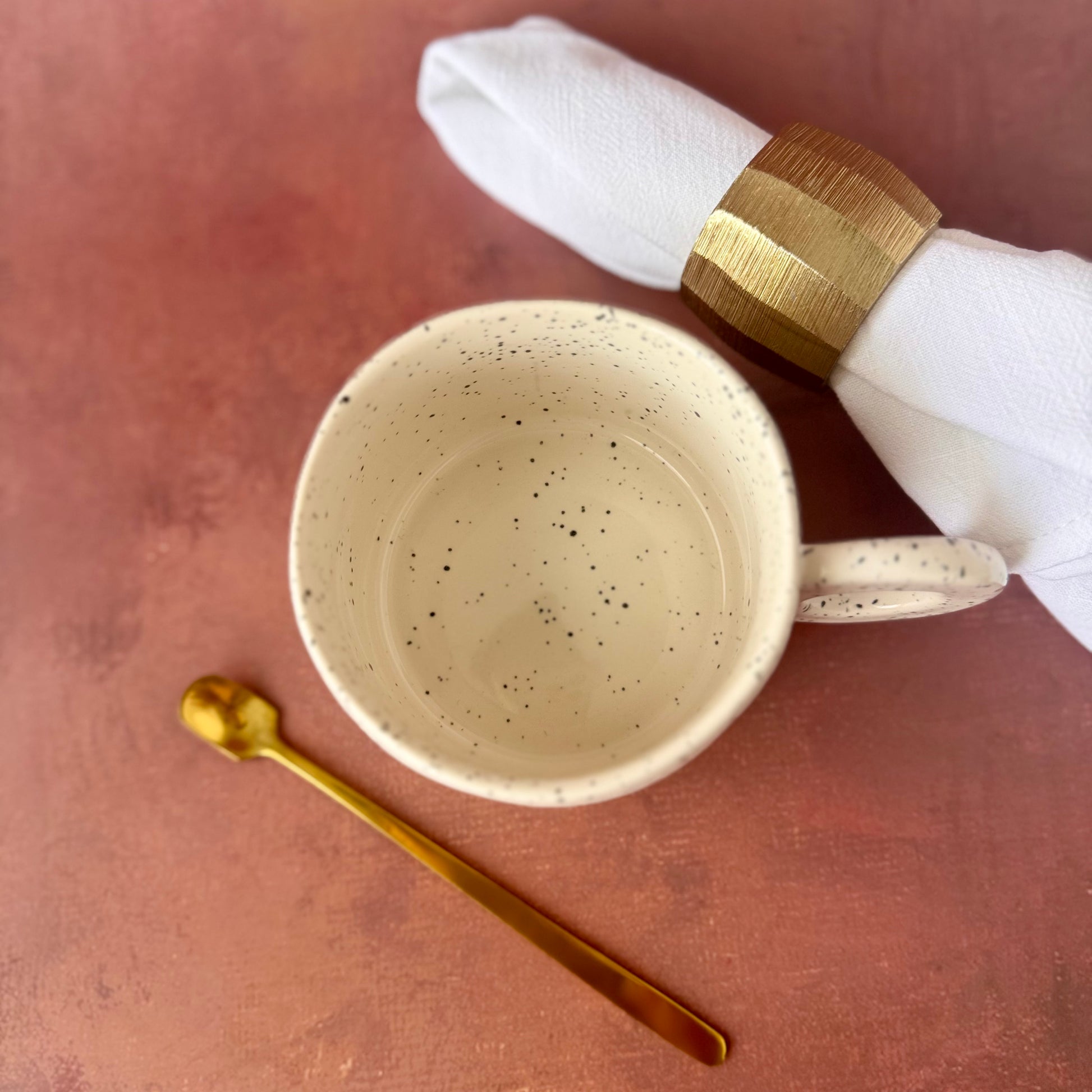 Ceramic white stylish mug with speckled texture, gold spoon, and white napkin with gold band on a brown surface