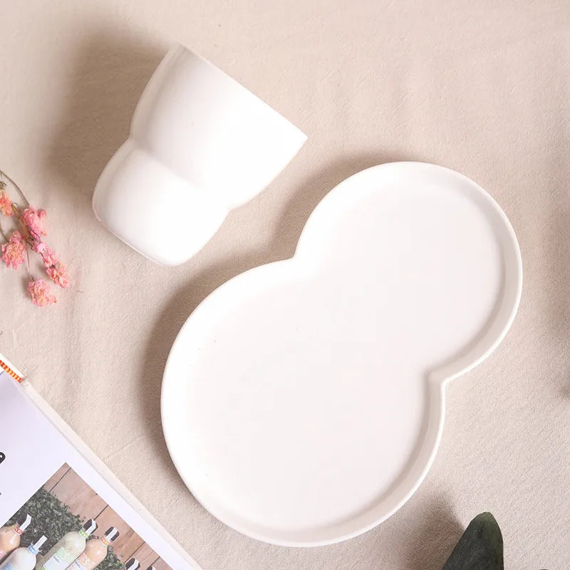 White ceramic round-shaped tray and cup on a beige surface with pink flowers and a magazine.