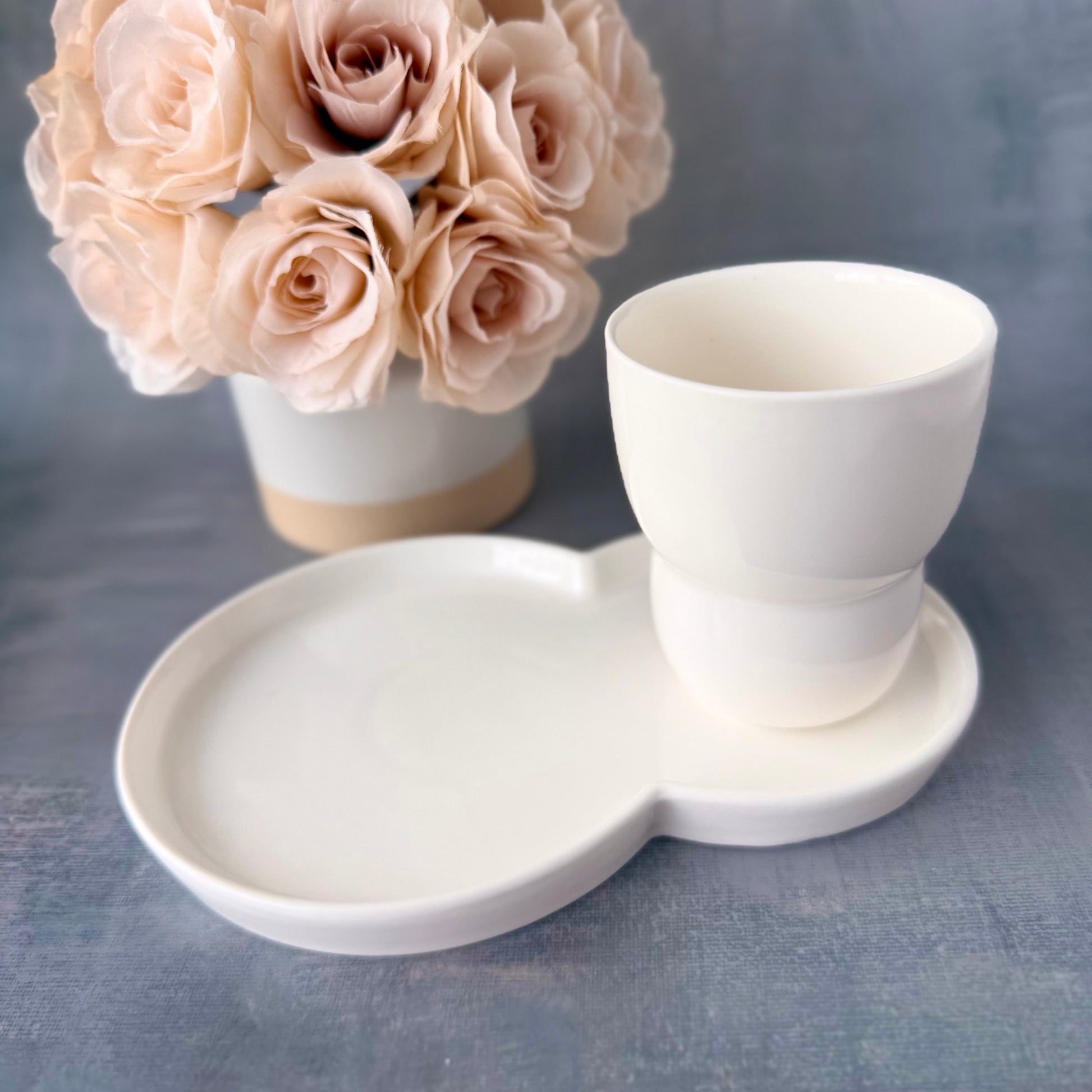 White ceramic cup and tray with a vase of beige roses on a gray background