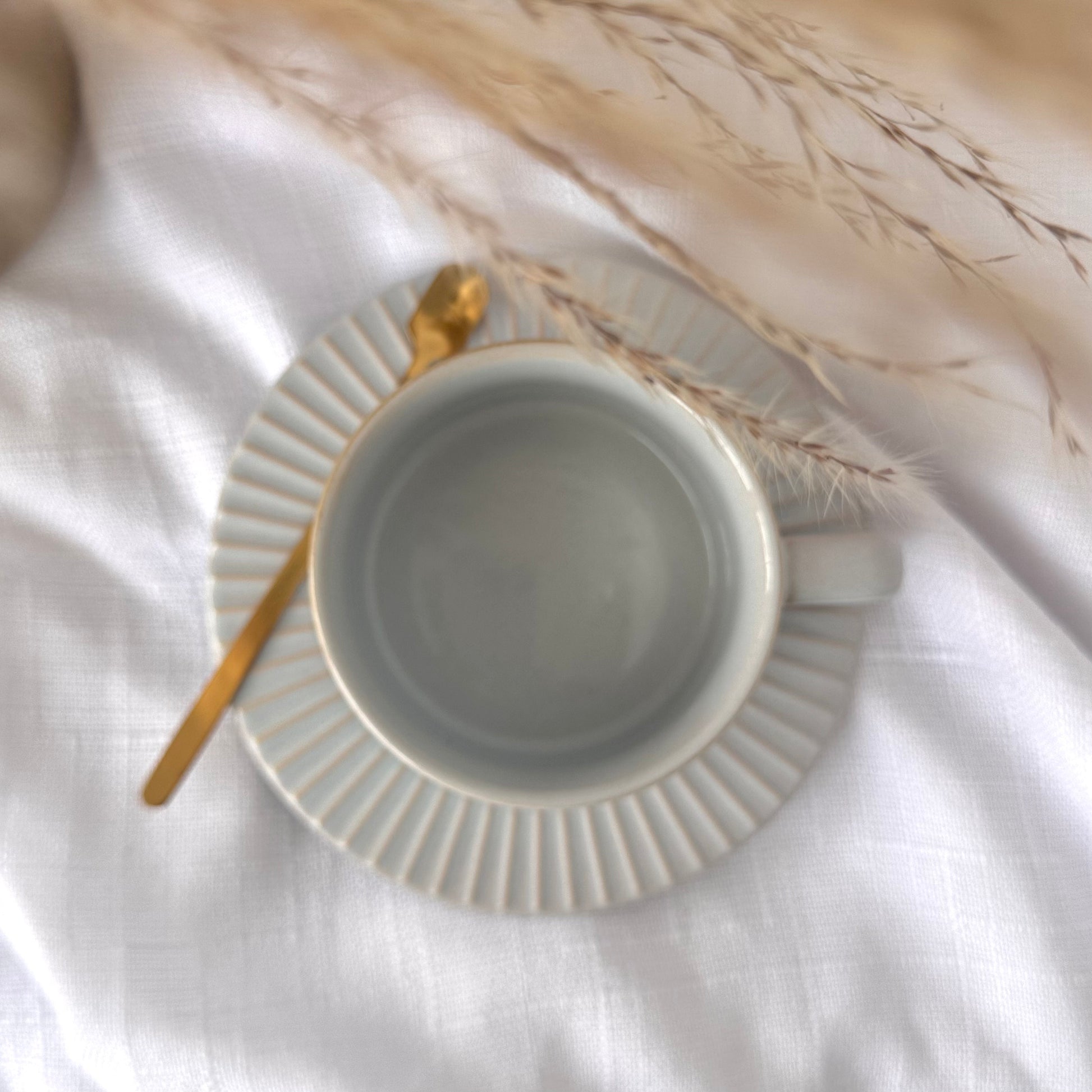 Calming light gray glaze with soft blue undertones ceramic cup with gold spoon on a white surface with dried plants