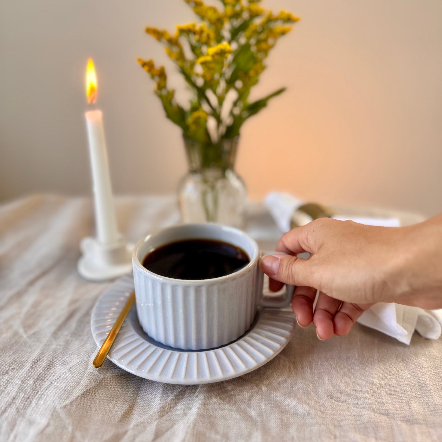 Person holding a calming light gray glaze with soft blue undertones mug of coffee on a table with a lit candle and flowers in the background.