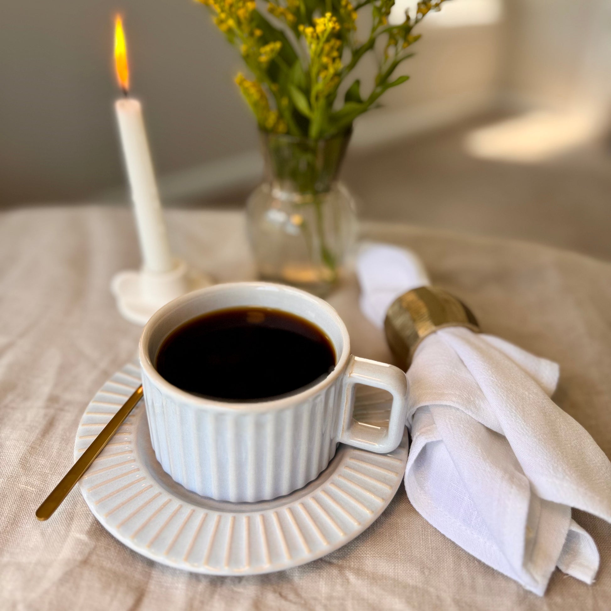 Calming light gray glaze with soft blue undertones mug with black coffee on a beige tablecloth, next to a lit candle and a vase with flowers.